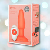A box of the B-Vibe Rimming Plug 2 Vibrating Butt Plug - Orange by B-Vibe, featuring peach packaging with product and remote displayed, rimming sensation highlights, and award icons. Blurred pastel lights form the background.
